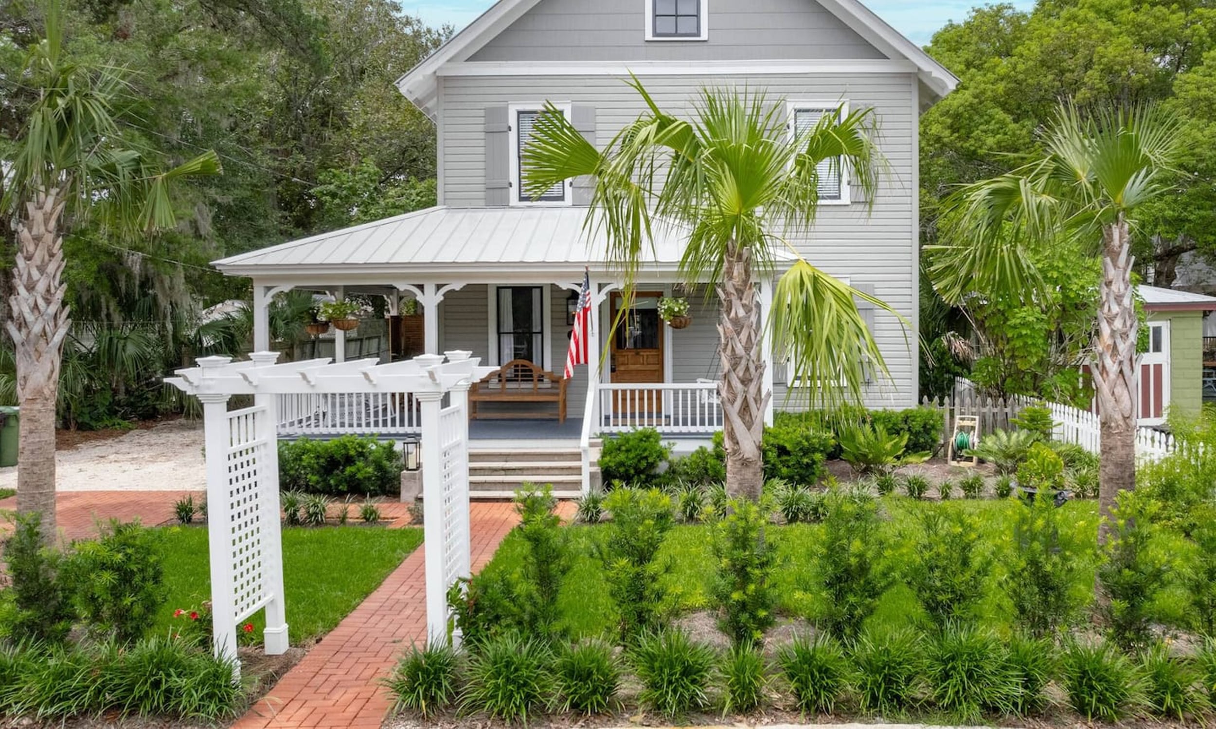 Exterior of home rental by Palm Row Hospitality featuring a pergola archway, palm trees, and a wraparound porch