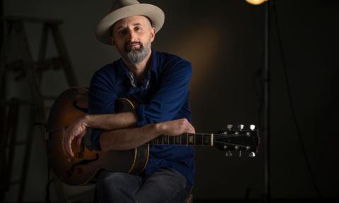 Seth Walker leans on his guitar and sits in front of a dark backdrop.