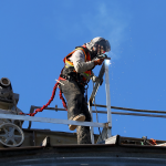A full color picture of a construction worker on a metal roof above the photographer. He is dressed fully in protective gear and is hooked to the roof with a safety chord as he welds. Sparks and smoke rise from his tools into the blue sky behind him.