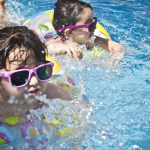 A full color image of two young dark haired girls in a swimming pool. They both use pink and yellow floaties.