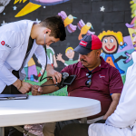 A full color image of two doctors caring for a patient (middle) both doctors are young men and their patient (wearing a hat) is an older man. 